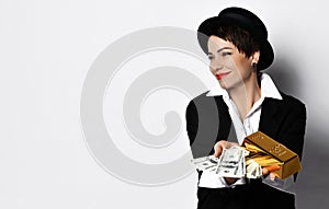 Smiling woman in hat and jacket demonstrates a fan of dollars and a bar of gold in her hands looks at free copy space