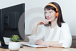 Smiling woman in headphones sitting at the table looking at the monitor screen, watching webinar or training course