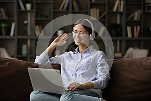 Smiling woman in headphones waving hand at webcam, using laptop