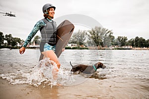 Smiling woman in helmet and wetsuit walks on water and dog runs nearby