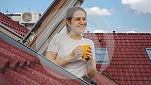 Smiling woman in her morning ritual as she sips tea or coffee at an attic window, basking in the view of red-tiled roofs