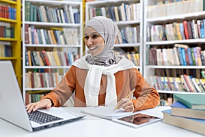 Smiling woman in hijab studying at library with laptop