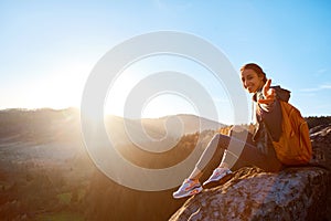 Smiling woman hiker sits on edge of cliff against background of sunrise