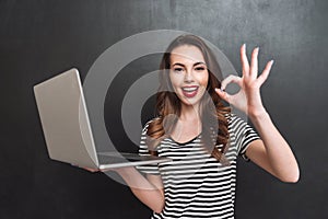 Smiling woman holding laptop computer and showing ok sign
