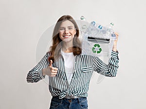 Smiling woman holding recycle bin with plastic bottles and showing thumb up