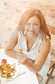 Smiling woman holds glass of white wine sitting at table On the sandy beach outdoors. Happy middle aged woman have date