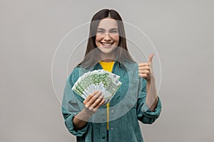 Smiling woman holds many euro banknotes and showing thumb up to camera, satisfied of winning lottery