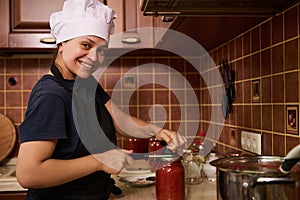 Smiling woman housewife in white chef hat and apron, using seamer, closes lids of jars with freshly canned tomato sauce