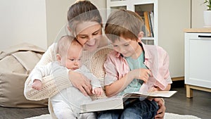 Smiling woman hugging her baby and older son while reading story book at living room. Parenting, children happiness and