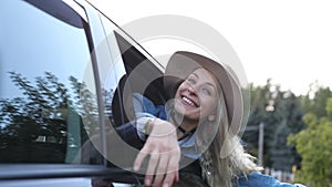 Smiling woman joyfully leave city sticking head out of open car window, from below view. Young blonde lady in hat nod