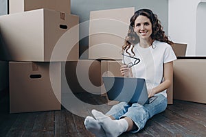 Smiling woman with laptop sitting on floor near cardboard boxes on moving day to new house