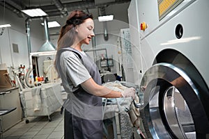 Smiling woman laundry service operator putting clothes into dry cleaning machine