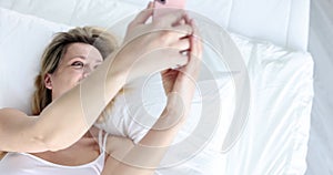 Smiling woman lies on bed and looks at smartphone screen