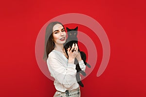 Smiling woman in light casual clothes stands on a red background with a black cat in her hands, looks into the camera and smiles.