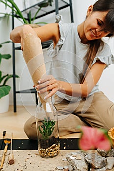 Smiling woman in living room planting inside glass bottle, using paper cone