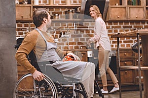 smiling woman looking at disabled husband in wheelchair holding vegetables while cooking dinner together