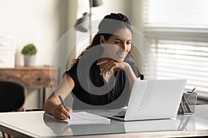 Smiling woman looking at laptop screen, taking notes, studying online