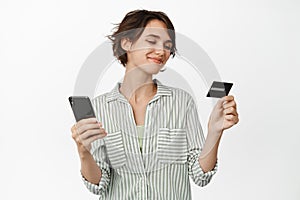 Smiling woman looking satisfied at credit card, holding mobile phone, paying online, making purchase, standing against