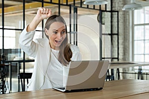 Smiling woman looks at the laptop screen. An excited business woman raises her hands up, has received a good business