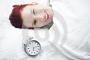 Smiling woman lying in bed with alarm clock on pillow