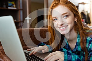 Smiling woman lying on brown sofa and using laptop