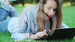 Smiling woman lying on green grass and typing on laptop in summer park