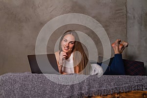 Smiling woman lying on her stomach in bed with laptop in front of her and looking into camera