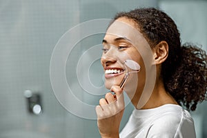 Smiling Woman making face massage using roller with quartz in bathroom. Natural cosmetics concept photo