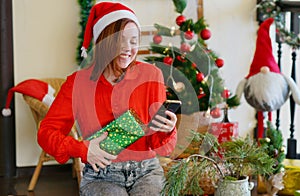 Smiling woman with a mobile Phone in hands and present. Christmas tree, lights. New year atmosphere.