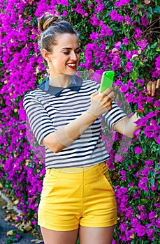 Smiling woman near flowers bed taking photo with cellphone