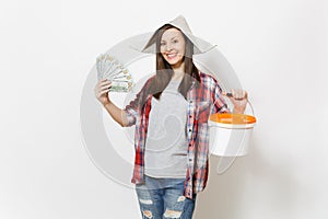 Smiling woman in newspaper hat holding bundle of dollars, cash money and empty paint bucket with copy space isolated on