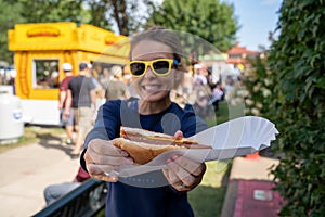 Smiling woman at an outdoor fair holds up a half-eaten footlong hot dog. Focus on the hot dog, girl intentionally blurred