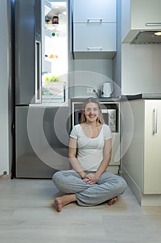 Smiling woman in pajamas sitting on floor at kitchen next to ope