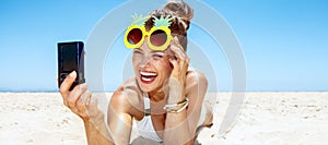 Smiling woman in pineapple glasses taking selfie at sandy beach