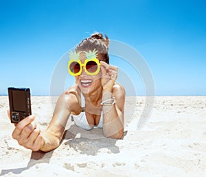 Smiling woman in pineapple glasses taking selfie at sandy beach