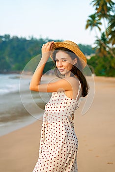 Smiling woman in polka dot dress and straw hat stands by tropical beach at sunrise. Happy female enjoys early morning on