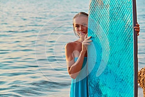 Smiling woman posing with surfboard an seashore. Happy girl surfer in blue dress. Summertime and time to adventure idea