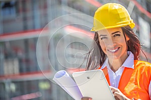 Smiling woman in PPE holds a tablet and a blueprint with a construction site in the background