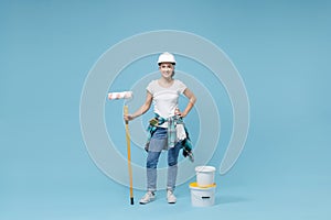 Smiling woman in protective helmet hardhat, glasses hold paint bucket, roller for wall painting isolated on blue