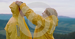 Smiling woman putting on hood of yellow rain coat on man standing on mountain peak. Side view portrait of loving mature