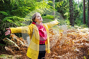 Smiling woman with raised arms in bright yellow raincoat enjoying life and freedom in autumn forest in nature. Feeling