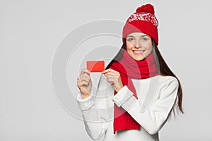 Smiling woman showing blank credit card, winter concept. Happy girl in red hat and scarf holding card, isolated over gray backgrou