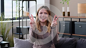 Smiling Woman Showing Victory Sign Sitting On Sofa At Home