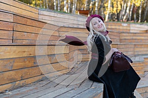 Smiling woman sits on a bench of a summer theater, wooden in a burgundy coat and biret, in the fall against a background