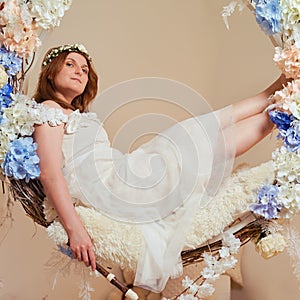 Smiling woman sits with a block flute on a swing in flowers. Studio portrait of happy female flutist in pink dress