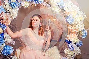 Smiling woman sits with a flute on a swing in flowers. Studio portrait of happy woman musician in pink dress