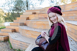 A smiling woman sits on a summer theater bench, made of wood in a burgundy coat and biret, an adult looks at the camera