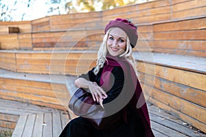 A smiling woman sits on a summer theater bench, made of wood in a burgundy coat and biret, an adult smiles at the camera
