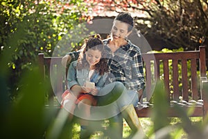 Smiling woman sitting with arm around daughter using mobile phone on wooden bench