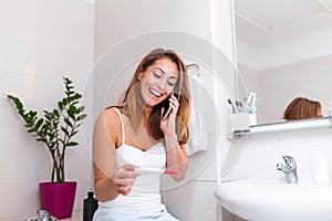 Smiling woman sitting in bathroom and holding positive pregnancy test and talking on her smart phone with the father to give him